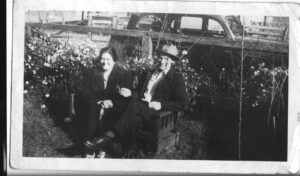 Two men sitting on a bench in front of an old car.