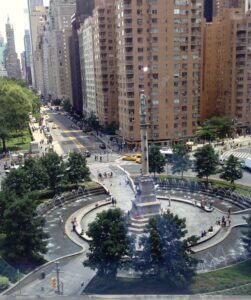 A view of the city from above shows a fountain, trees and buildings.